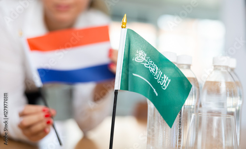 Little flag of Saudi Arabia on table with bottles of water and flag of Netherlands put next to it by positive young woman in meeting room photo