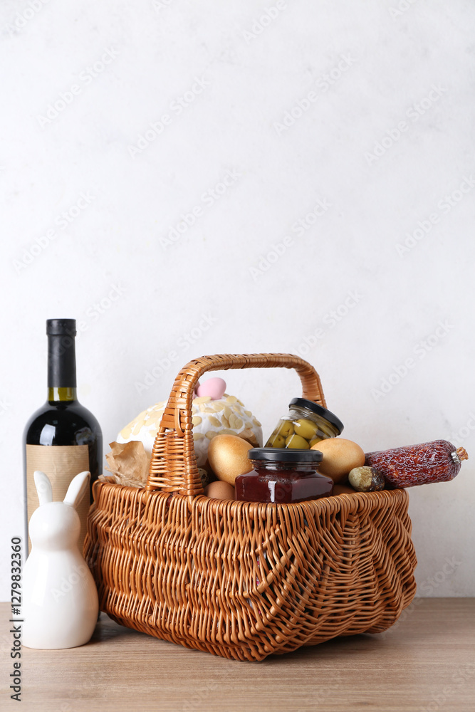 Naklejka premium Basket with Easter eggs, jars, sausage and bottle of wine on wooden table against white background