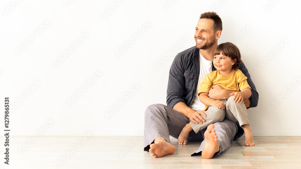 Great family ad. Happy caucasian father carrying his little son, sitting together on floor and looking aside at empty space, white wall background, mockup for text Great family ad. Happy caucasian father carrying his little son, sitting together on floor and looking aside at empty space, white wall background, mockup for text