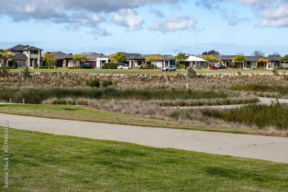 A suburban neighborhood in Melbourne, Australia, featuring a row of modern single-story houses with a green park with a walking path, and natural wetlands with newly planted vegetation. A suburban neighborhood in Melbourne, Australia, featuring a row of modern single-story houses with a green park with a walking path, and natural wetlands with newly planted vegetation.