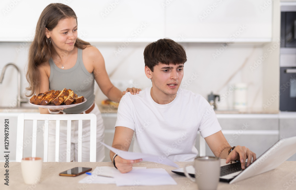 Young woman offering piece of pie to young guy working at laptop in kitchen Young woman offering piece of pie to young guy working at laptop in kitchen