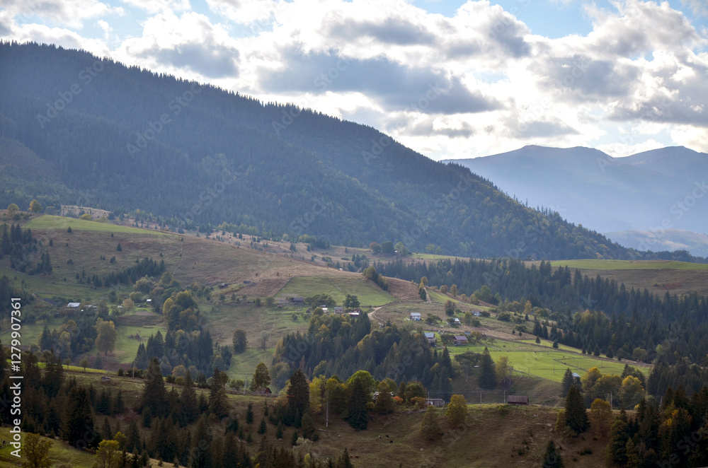 Fototapeta premium Idyllic rural scenery featuring green rolling hills spotted with small houses and farm structures, surrounded by lush greenery and distant forested mountains under a cloudy sky. Carpathians, Ukraine