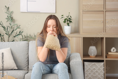 Emotional young woman with paper bag having panic attack on sofa at home