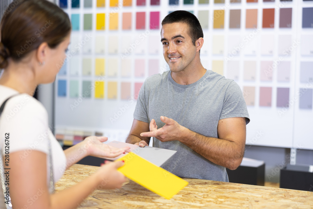 Male salesperson in paint and varnish department is assisting female customer by showcasing product samples and providing guidance through product catalog at counter. Male salesperson in paint and varnish department is assisting female customer by showcasing product samples and providing guidance through product catalog at counter.