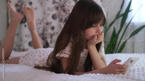 Girl is looking at her smartphone, lying in bed at her leisure. Teenage girl is lying on bed and playing video games on her phone. Concept of the problem of children's gadgets.