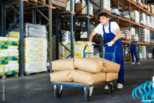 фотографія In warehouse rack area of store, stevedore loader young man pushes and carries l