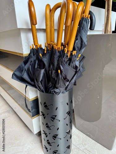 A black umbrella stand with decorative cut-out umbrella patterns holds multiple wooden-handle black umbrellas in an indoor setting. The polished floor and modern decor add to the aesthetic.
