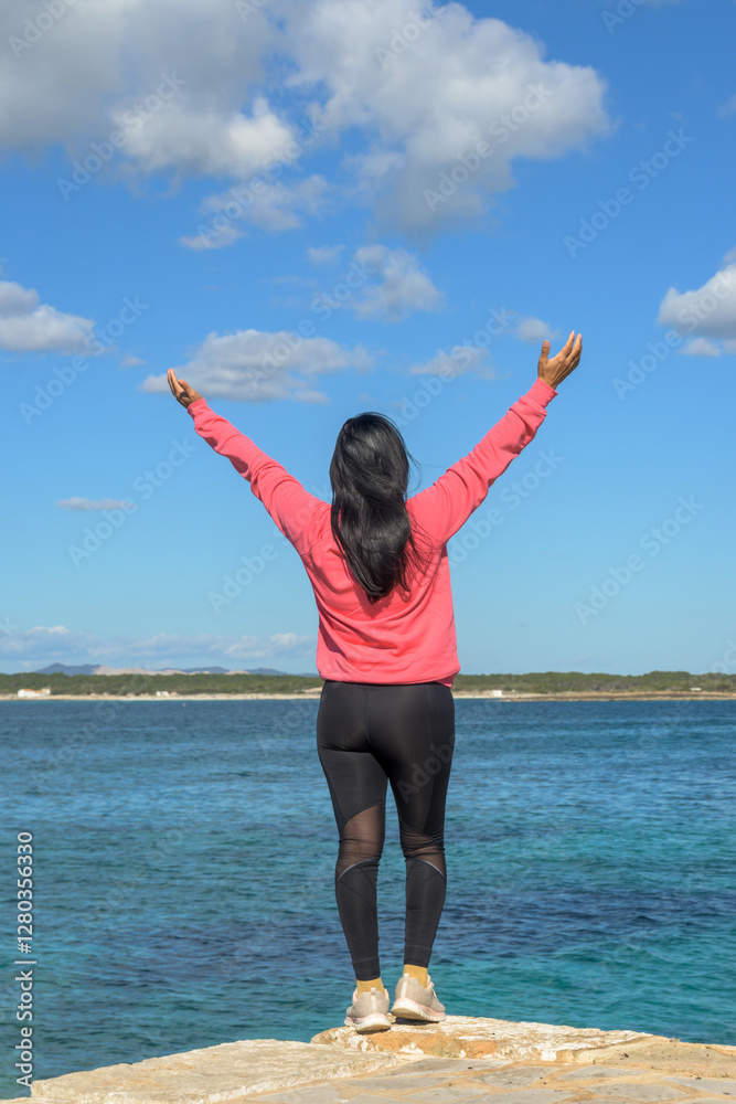 Woman facing the sea with arms raised, wearing pink and black Woman facing the sea with arms raised, wearing pink and black
