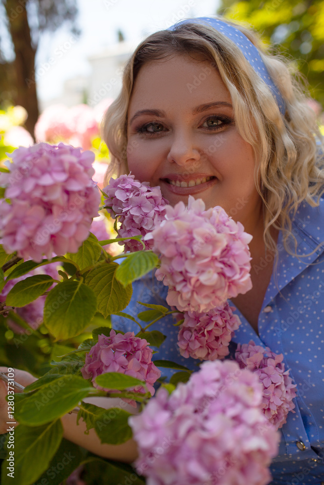 Portrait of a beautiful fat girl in a pale blue dress, against the background of a wall of live growing pink gartenias. The plus size woman enjoys the beauty of flowers Portrait of a beautiful fat girl in a pale blue dress, against the background of a wall of live growing pink gartenias. The plus size woman enjoys the beauty of flowers