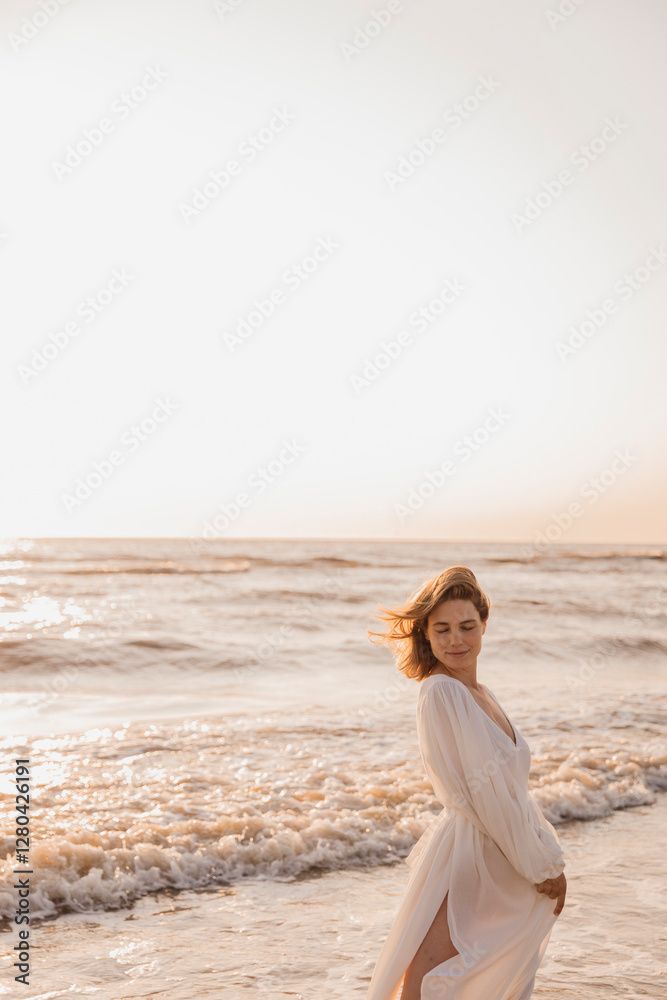 Woman in a flowing white dress standing in ocean waves at sunset