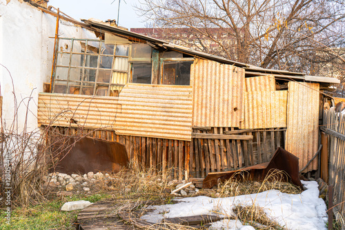 A weathered shack constructed from corrugated metal sheets, old wooden planks, and broken windows. The structure appears aged and abandoned, surrounded by dry grass, rocks, and patches of snow.