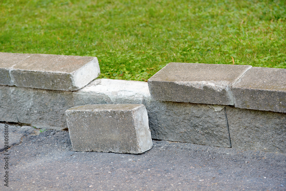 Damaged concrete curb separating grass and asphalt, with one block displaced. Broken curb stone lying on asphalt, need for repair and maintenance in an urban infrastructure setting. Selective focus