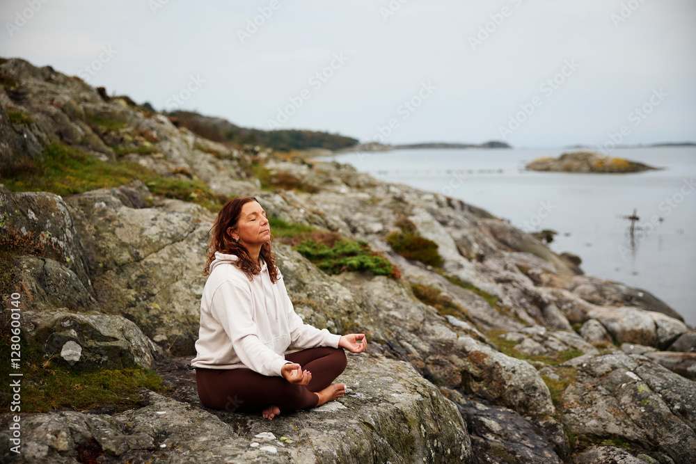 Full length of woman meditating while sitting on rocks by sea Full length of woman meditating while sitting on rocks by sea