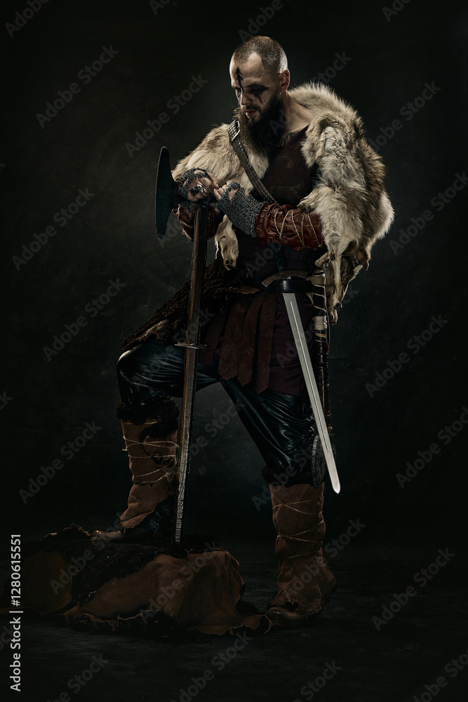 Obraz premium Viking warrior in rugged fur cloak, with axe and sword, standing in dramatic lighting against dark background. Concept of warriors bravery, medieval strength, Norse legend, historical portrait