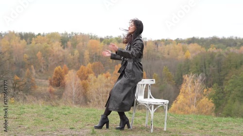 Autumn landscape with gothic woman on chair. Stylish woman and chair in autumn landscape outdoor. Fall nature landscape and girl. Central park in New York. Girl in fall trench. Loneliness concept