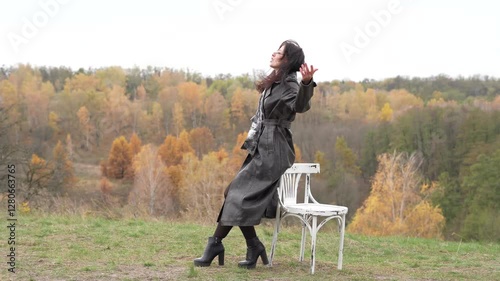 Autumn landscape with gothic woman on chair. Stylish woman and chair in autumn landscape outdoor. Fall nature landscape and girl. Central park in New York. Girl in fall trench. In a field.