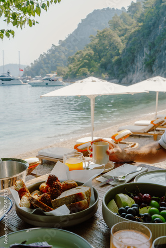 Fototapeta Naklejka Na Ścianę i Meble -  A picturesque seaside breakfast setup in Fethiye, Türkiye, featuring fresh fruits, cheeses, bread, and olives on a rustic wooden table with ocean views, white umbrellas, and yachts in the background