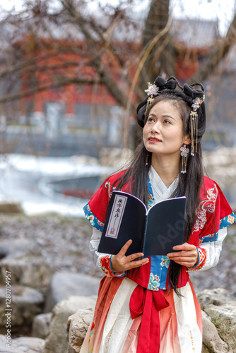 A young Asian woman in a traditional red Chinese costume walks in the park, reads national haiku poetry. Celebrating Chinese New Year