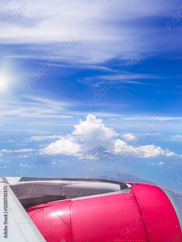 Aerial view from airplane window showing a red engine, blue sky, white clouds, and coastline below. The horizon stretches far, blending ocean and land in a breathtaking travel scene.