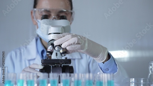 A scientist woman, wearing a lab coat, mask, white gloves and safety glasses, is adjusting a microscope surrounded by test tubes in laboratory