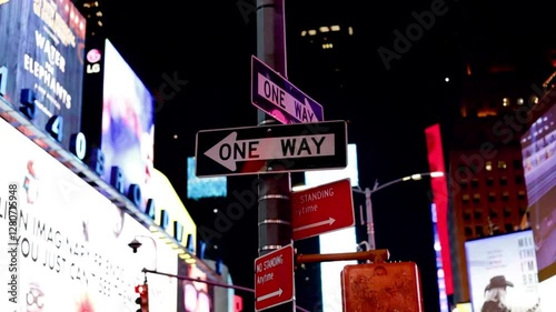 A cluster of illuminated street signs, including a one-way sign, stands against the bright lights and towering billboards of Times Square at night, capturing the energy and vibrancy of New York City