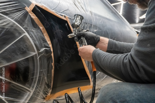 Painting technician applying a coat of paint on the fender of the car.  Local repair of car paintwork. A painter with a spray gun paints a black car. Spray gun in the painter's hand. 