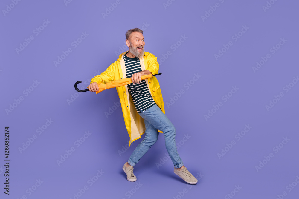 Joyful senior man in a yellow raincoat holding a closed umbrella playfully against a vibrant purple background Joyful senior man in a yellow raincoat holding a closed umbrella playfully against a vibrant purple background