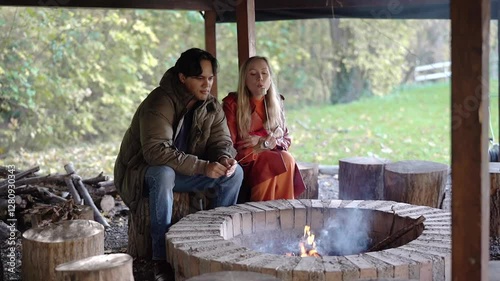 A young couple sits by a rustic fire pit, roasting marshmallows and enjoying a peaceful outdoor moment. The man is focused on toasting his marshmallow, while the woman smiles warmly, holding hers. A p