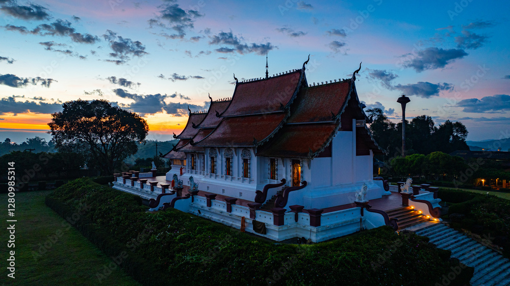 Obraz premium Aerial view of a traditional temple surrounded by greenery under a vibrant sunrise sky. The golden and blue hues of twilight highlight the intricate architecture and peaceful ambiance of the scene