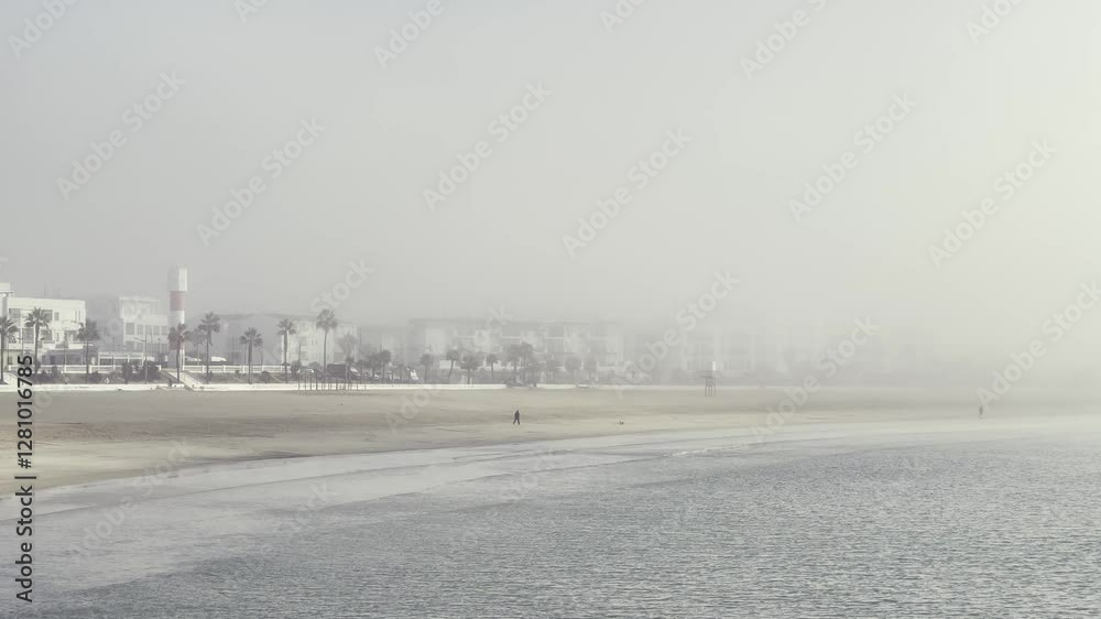 Barbate beach in Andalusia, Spain, is shrouded in fog on a misty morning, creating a mysterious and atmospheric scene with palm trees and buildings barely visible in the background Barbate beach in Andalusia, Spain, is shrouded in fog on a misty morning, creating a mysterious and atmospheric scene with palm trees and buildings barely visible in the background