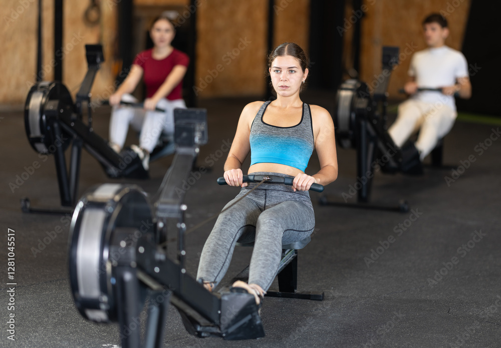 In gym, girl does exercises using rowing machine. Young group of athletes trains in gym. Physical education and sports, active healthy lifestyle. In gym, girl does exercises using rowing machine. Young group of athletes trains in gym. Physical education and sports, active healthy lifestyle.