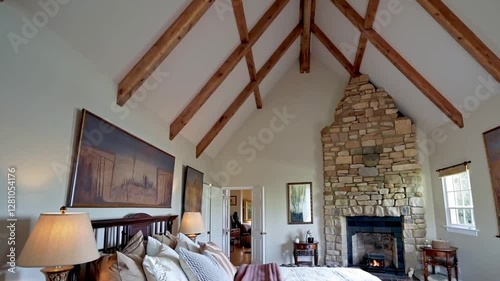 A bedroom with a vaulted ceiling, exposed wooden beams, and a rustic-modern vibe, highlighted by a stone fireplace and elegant textiles.