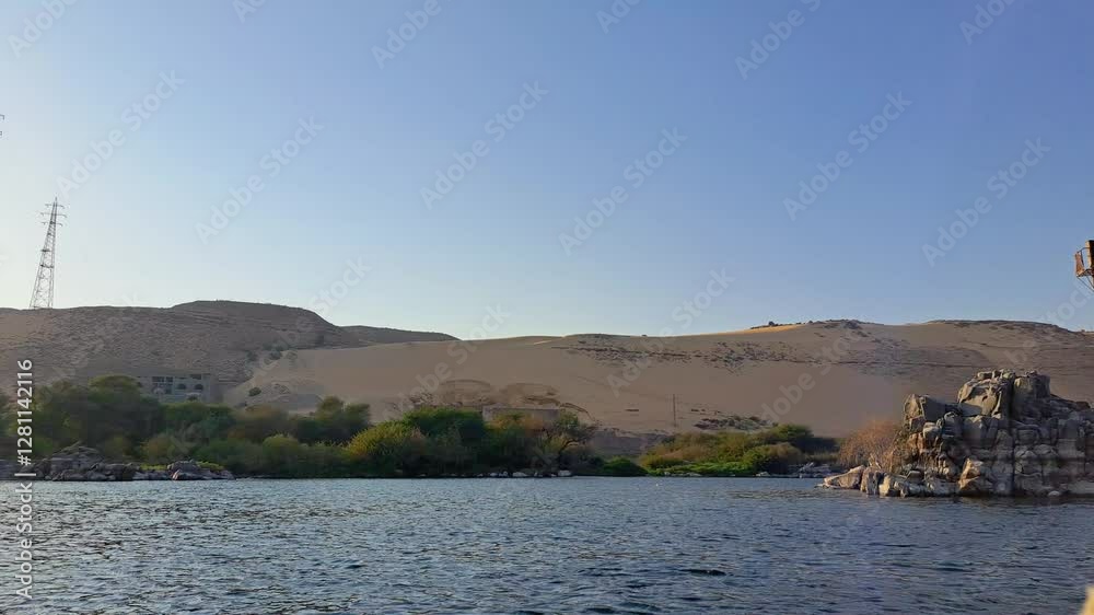 A serene view of the Nile River with lush greenery along the banks and towering sand dunes in the background. A power line tower stands prominently on the left side. Aswan, Egypt. A serene view of the Nile River with lush greenery along the banks and towering sand dunes in the background. A power line tower stands prominently on the left side. Aswan, Egypt.