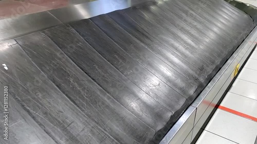 An empty conveyor belt awaits passengers in the baggage claim area of a busy airport. The polished surface reflects the surrounding terminal environment.