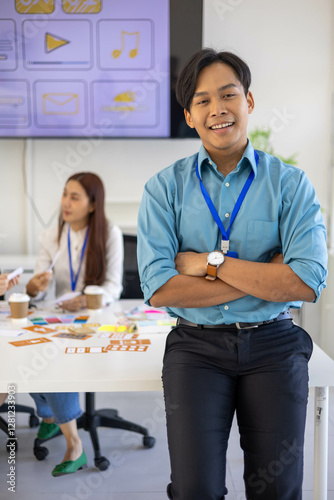A man in a blue shirt and black pants is smiling and standing in front of a white table. He is wearing a blue lanyard and a watch. Concept of professionalism and confidence

