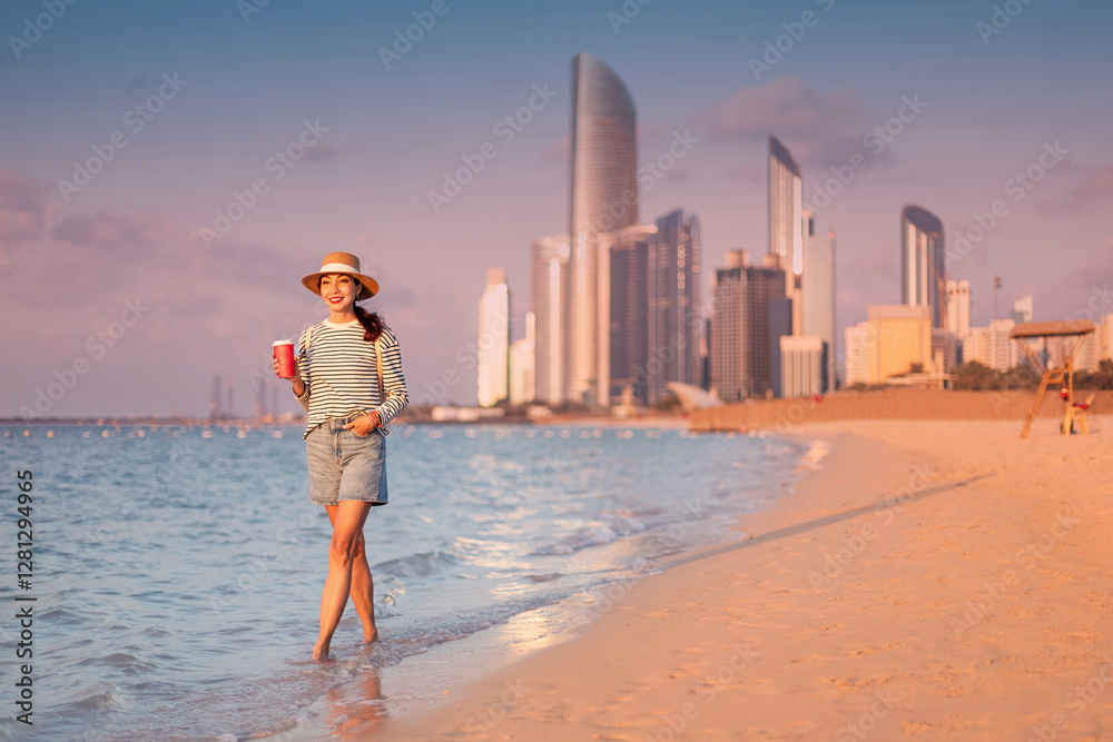 Young woman walking on beach holding beverage with Abu Dhabi skyline in background during sunset