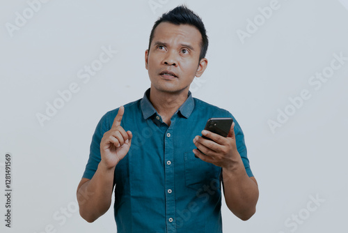 Asian man holding mobile phone with confused expression, isolated over white background.