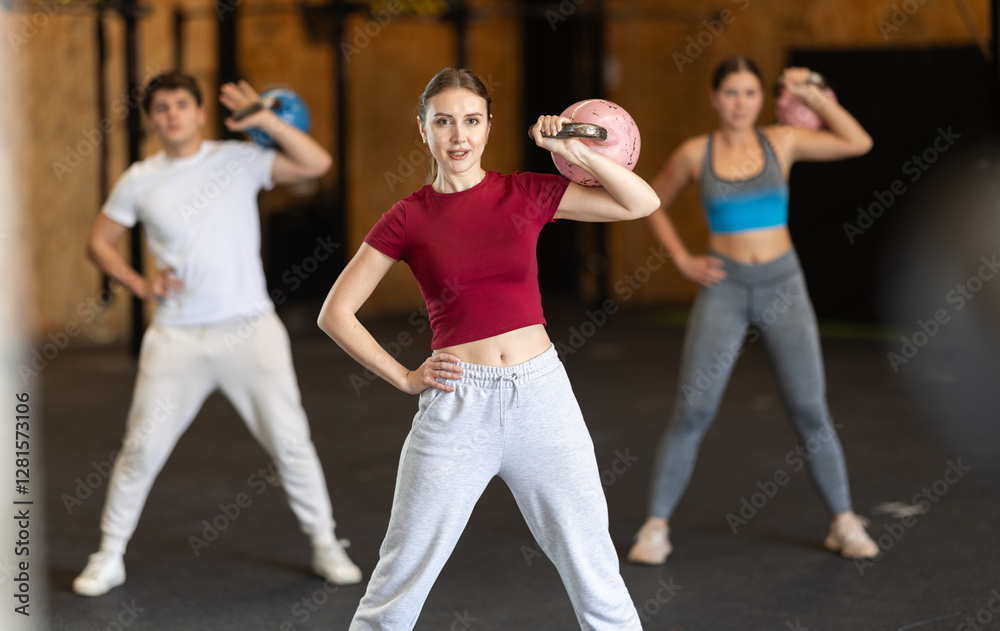 Naklejka premium During training, young woman teacher does exercises with kettlebell and leads group of athletes, gives sample of exercises. Leader raises and lowers kettlebell with one hand.