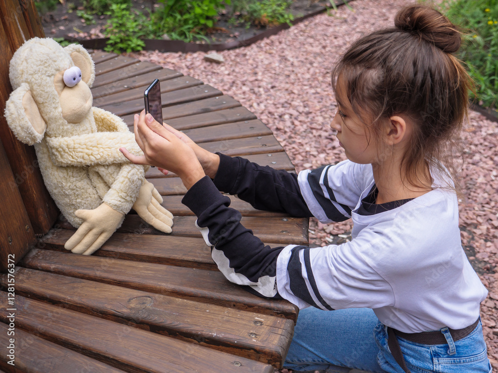 Cute girl with bun hairstyle holds smartphone and takes photo of her funny stuffed toy monkey with bulging eyes sitting on a wooden bench in a city park outdoors.