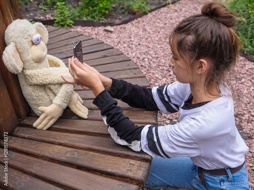 Obraz na płótnie Cute girl with bun hairstyle holds smartphone and takes photo of her funny stuffed toy monkey with bulging eyes sitting on a wooden bench in a city park outdoors. w salonie