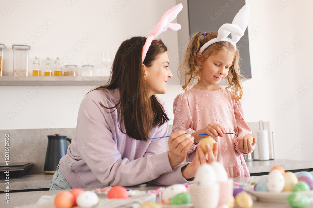 Smiling woman decorating Easter eggs with child girl 5-6 year old wearing bunny headbands at home kitchen table. Spring holidays. Smiling woman decorating Easter eggs with child girl 5-6 year old wearing bunny headbands at home kitchen table. Spring holidays.