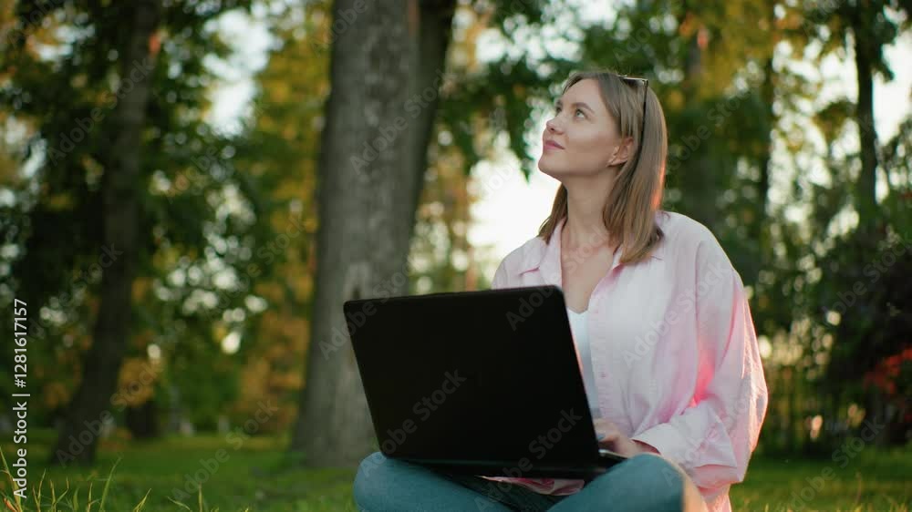 Young lady seated cross-legged on grassy field, working on laptop, looks up thoughtfully with a warm smile, background features trees and greenery, reflecting calm and productivity outdoors Young lady seated cross-legged on grassy field, working on laptop, looks up thoughtfully with a warm smile, background features trees and greenery, reflecting calm and productivity outdoors