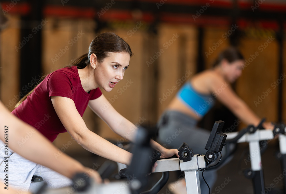 Focused girl works out with exercise bicycle during class at fitness club. Gym visitor performs exercise, creates load on muscles. Focused girl works out with exercise bicycle during class at fitness club. Gym visitor performs exercise, creates load on muscles.