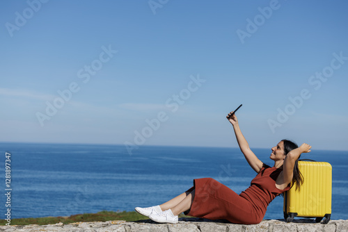 Smiling woman takes photos and selfies with her mobile phone with her yellow suitcase and the blue sky in the background, she is ready to start her summer trip. Concept of disconnection and enjoying.