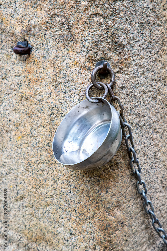 a metal bowl for drink water hanging from a chan at stone tap