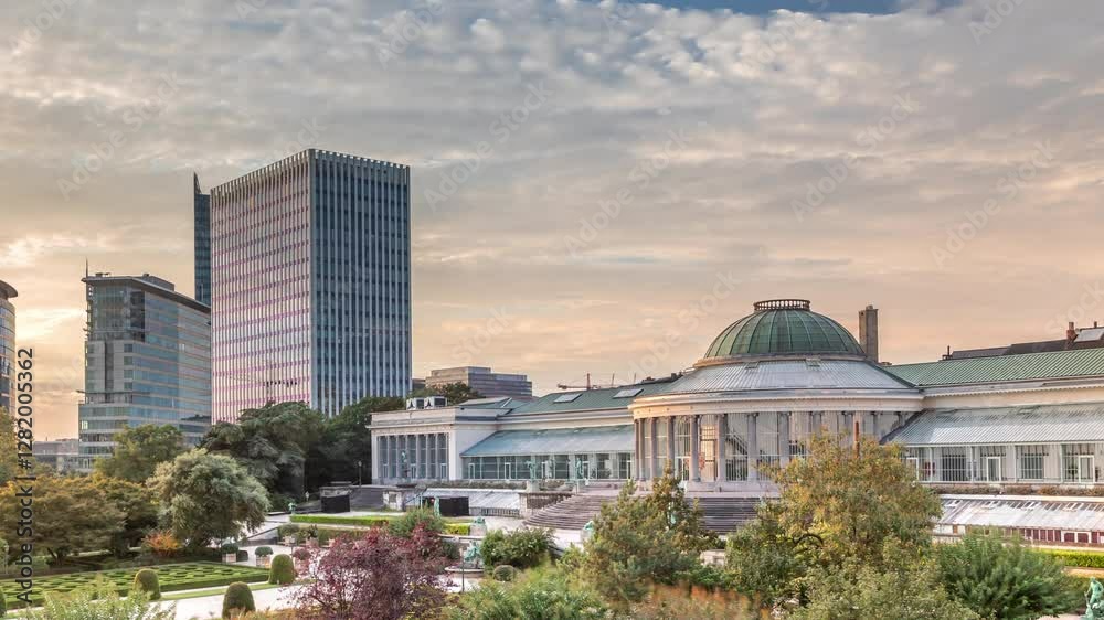 Aerial timelapse of Le Botanique and its orangery in Brussels, Belgium, surrounded by gardens and urban skyscrapers. Dome and walking people during sunset with orange clouds in the vibrant cityscape