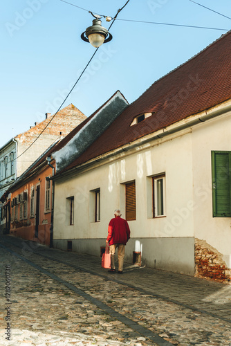 old man walking on medieval stone paved street with shopping bags in romania