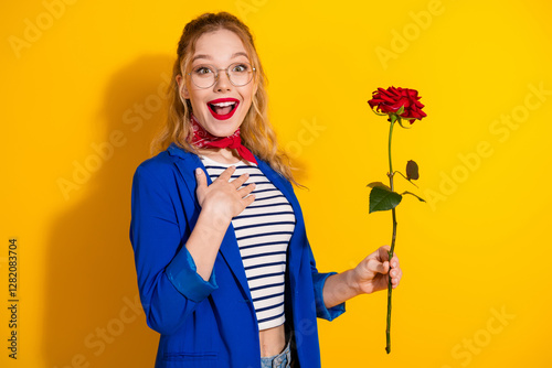 Young woman joyful over gift holding rose wearing glasses and blue jacket on a vibrant yellow background photo