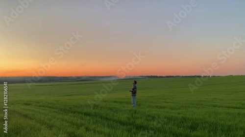 A guy with a cup of tea wanders through a green meadow against the background of an orange sky and enjoys the sunset