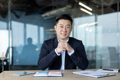 Portrait of a smiling and successful young Asian businessman and director sitting in a suit at his desk and looking confidently at the camera